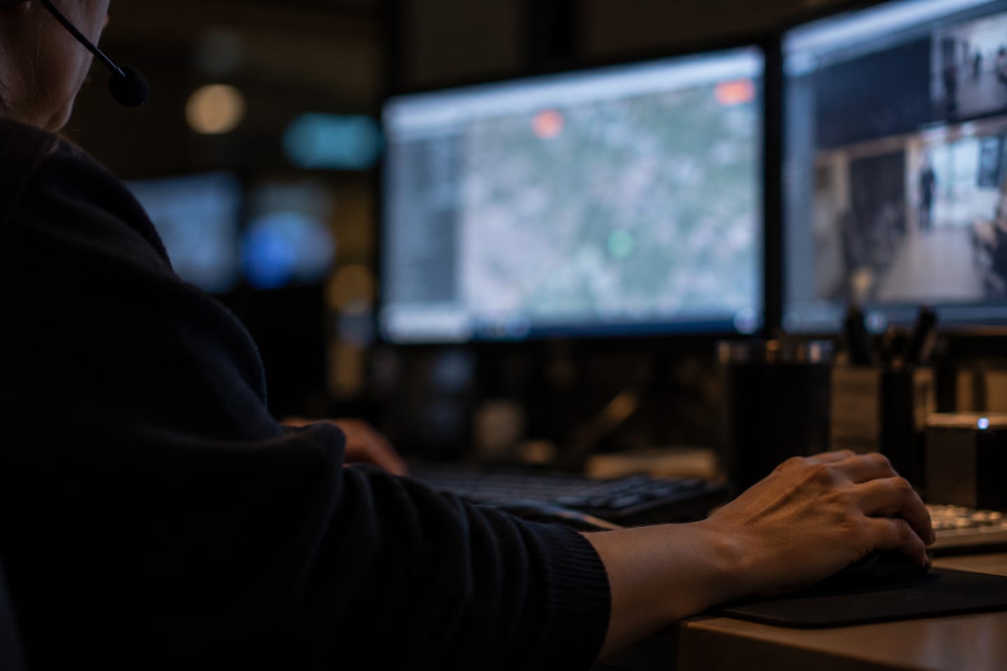 A monitoring operator's hand on a mouse in front of out-of-focus screens in a dark room